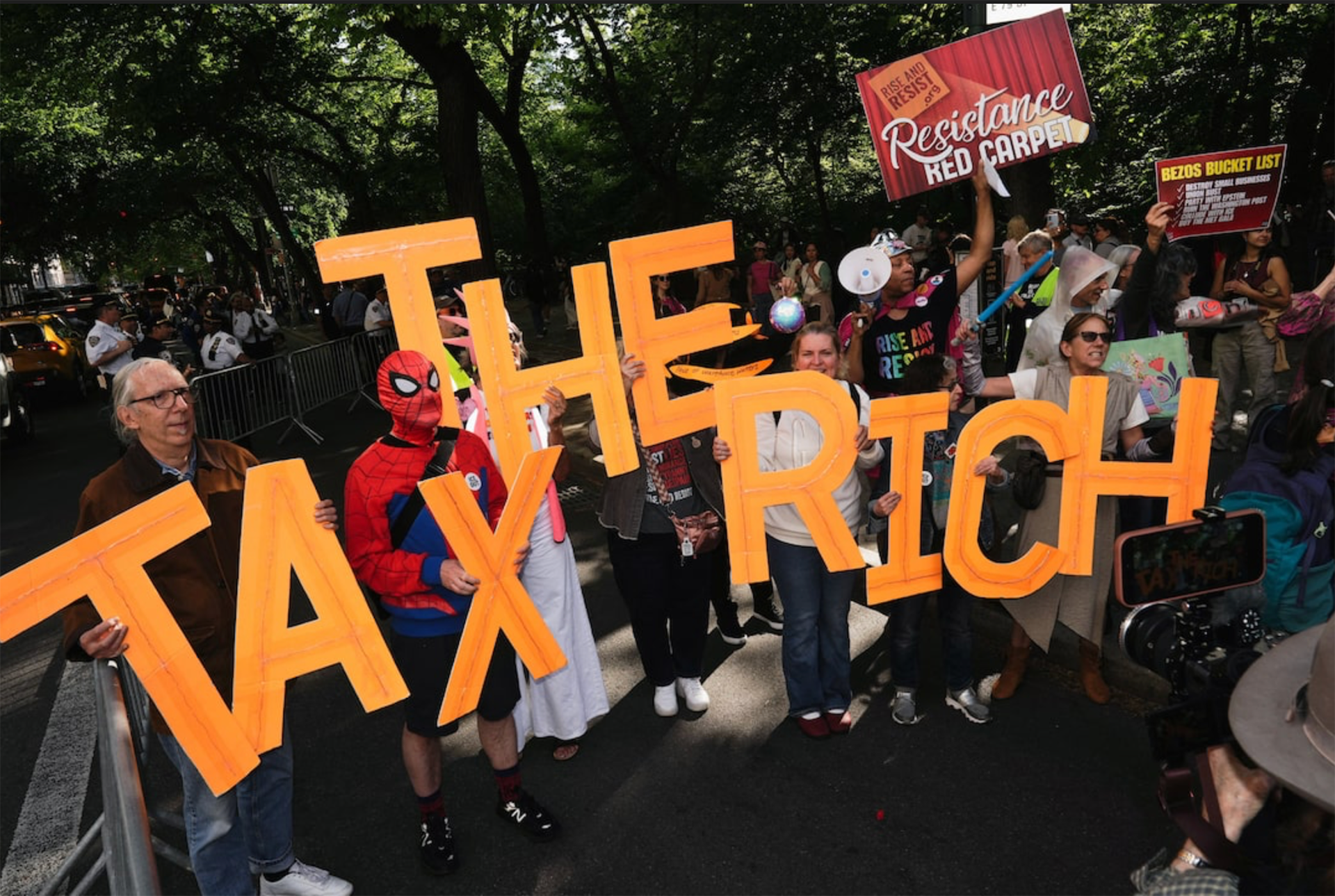 Tax the Rich Protesters hold large orange letters forming a banner across a street, with signs and a crowd behind a barricade in a park setting.